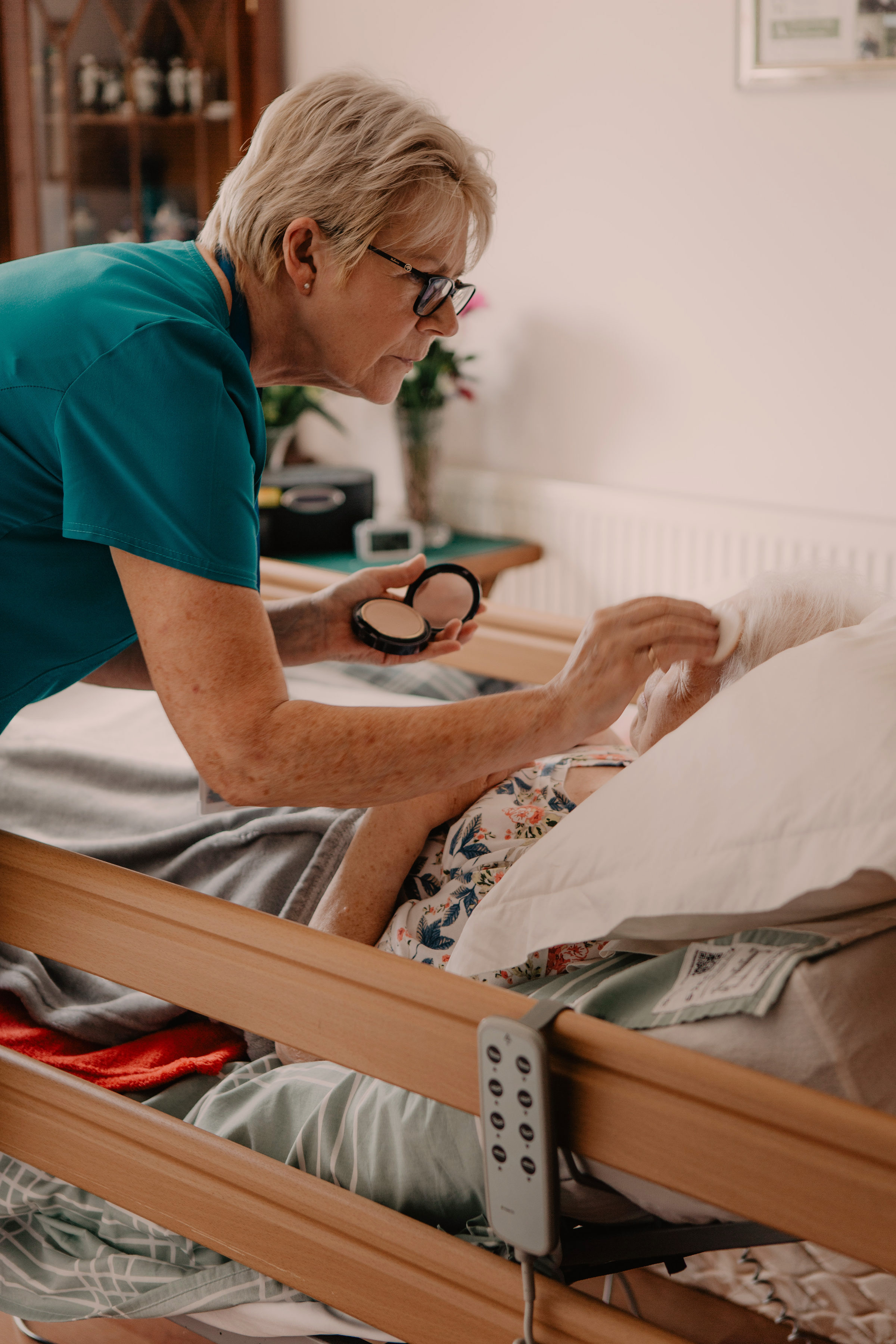 Female healthcare assistant, doing makeup from a compact for a female patient in a hospital bed at home