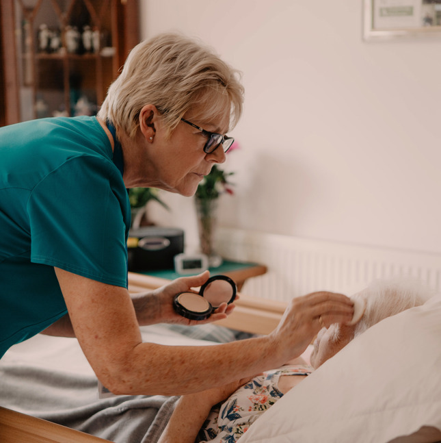 Female healthcare assistant, doing makeup from a compact for a female patient in a hospital bed at home
