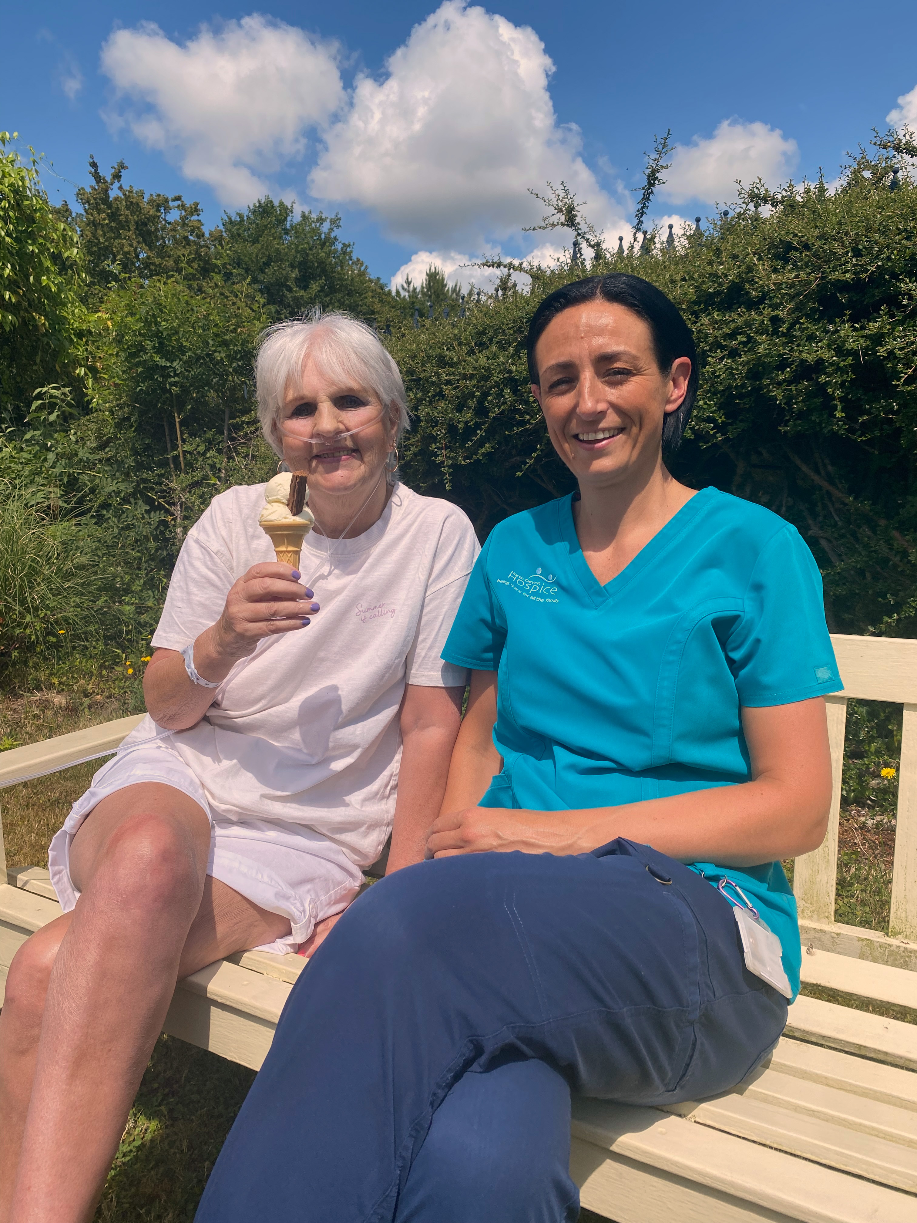 Female patient holding an icecream sat on a wooden bench with a female healthcare assistant, in the sunshine