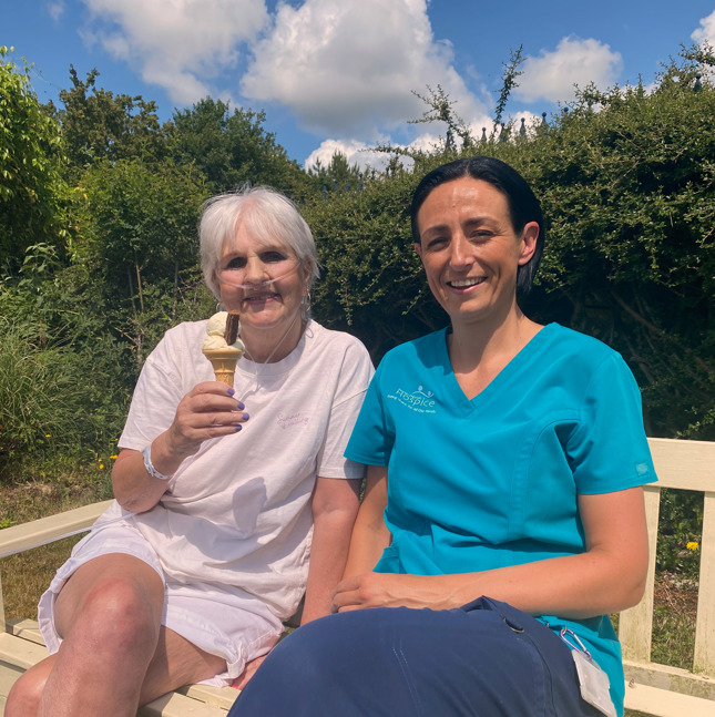 Female patient holding an icecream sat on a wooden bench with a female healthcare assistant, in the sunshine