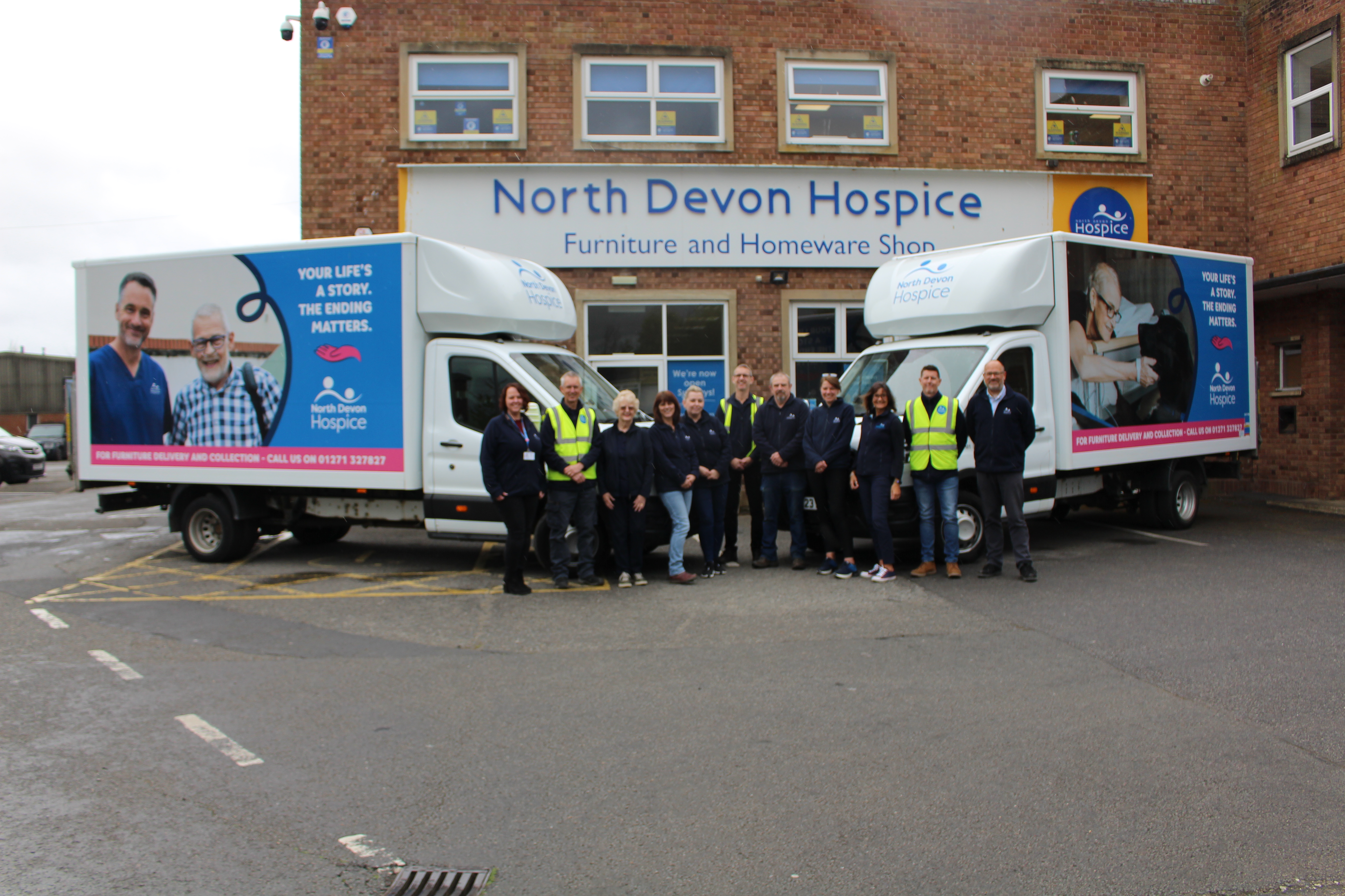 Team of charity shop workers stood in the centre of 2 hospice branded vans