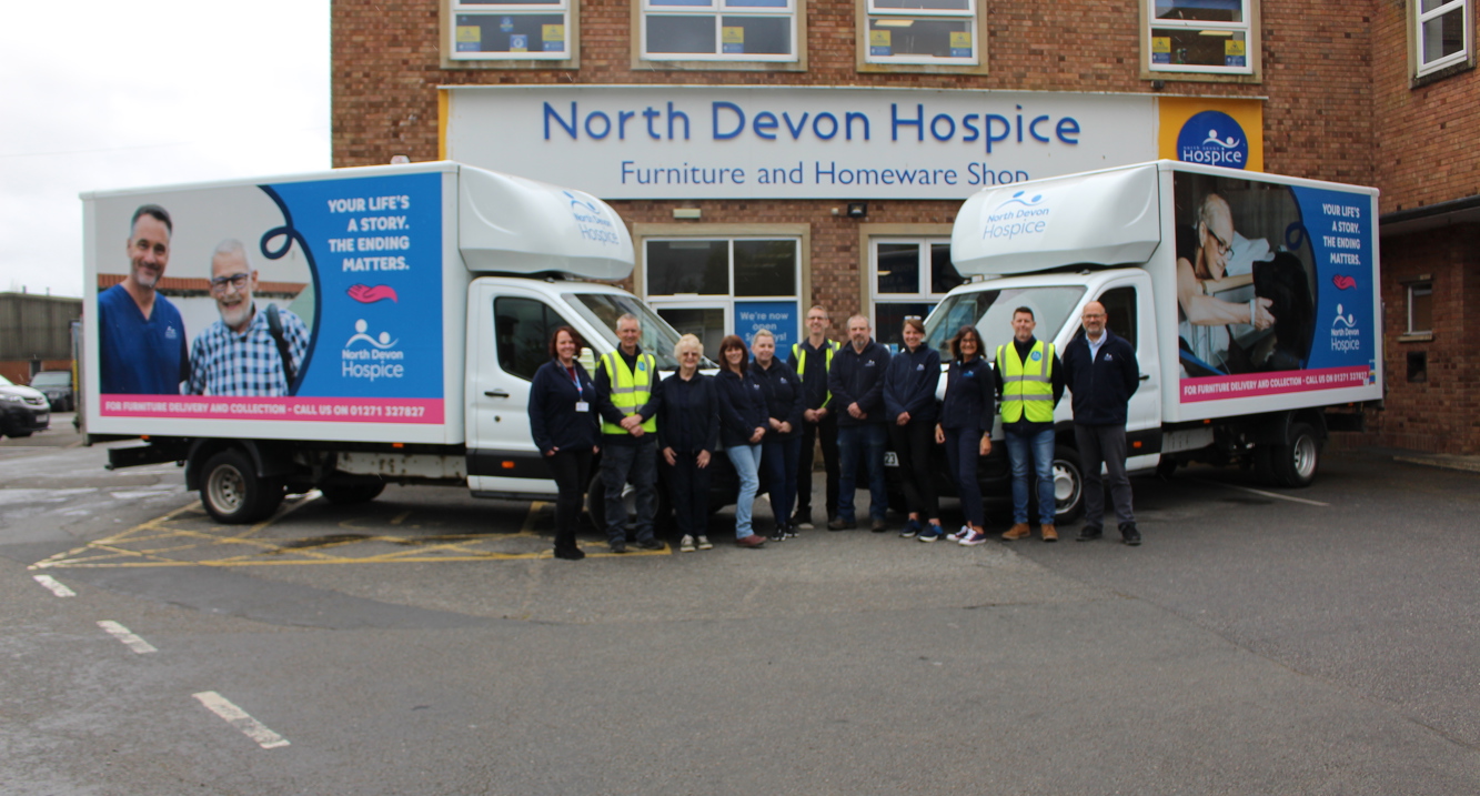 Team of charity shop workers stood in the centre of 2 hospice branded vans