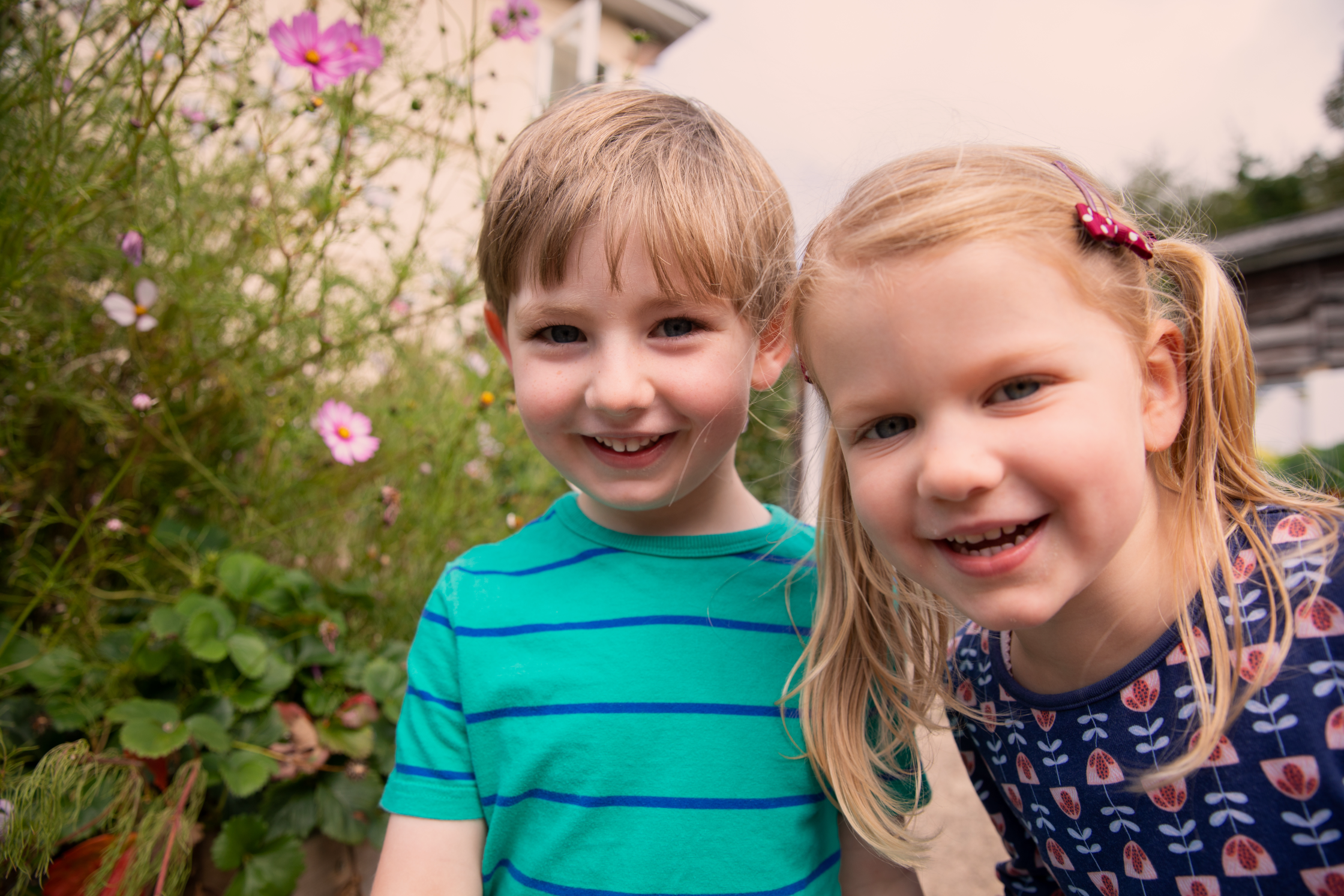 Small boy in green and blue striped t-shirt with a small girl with blonde pigtails, close to camera laughing, surrounded by flowers and plants
