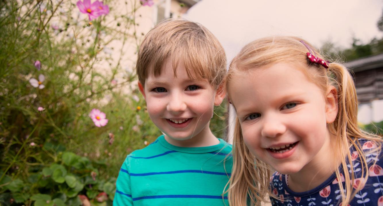Small boy in green and blue striped t-shirt with a small girl with blonde pigtails, close to camera laughing, surrounded by flowers and plants