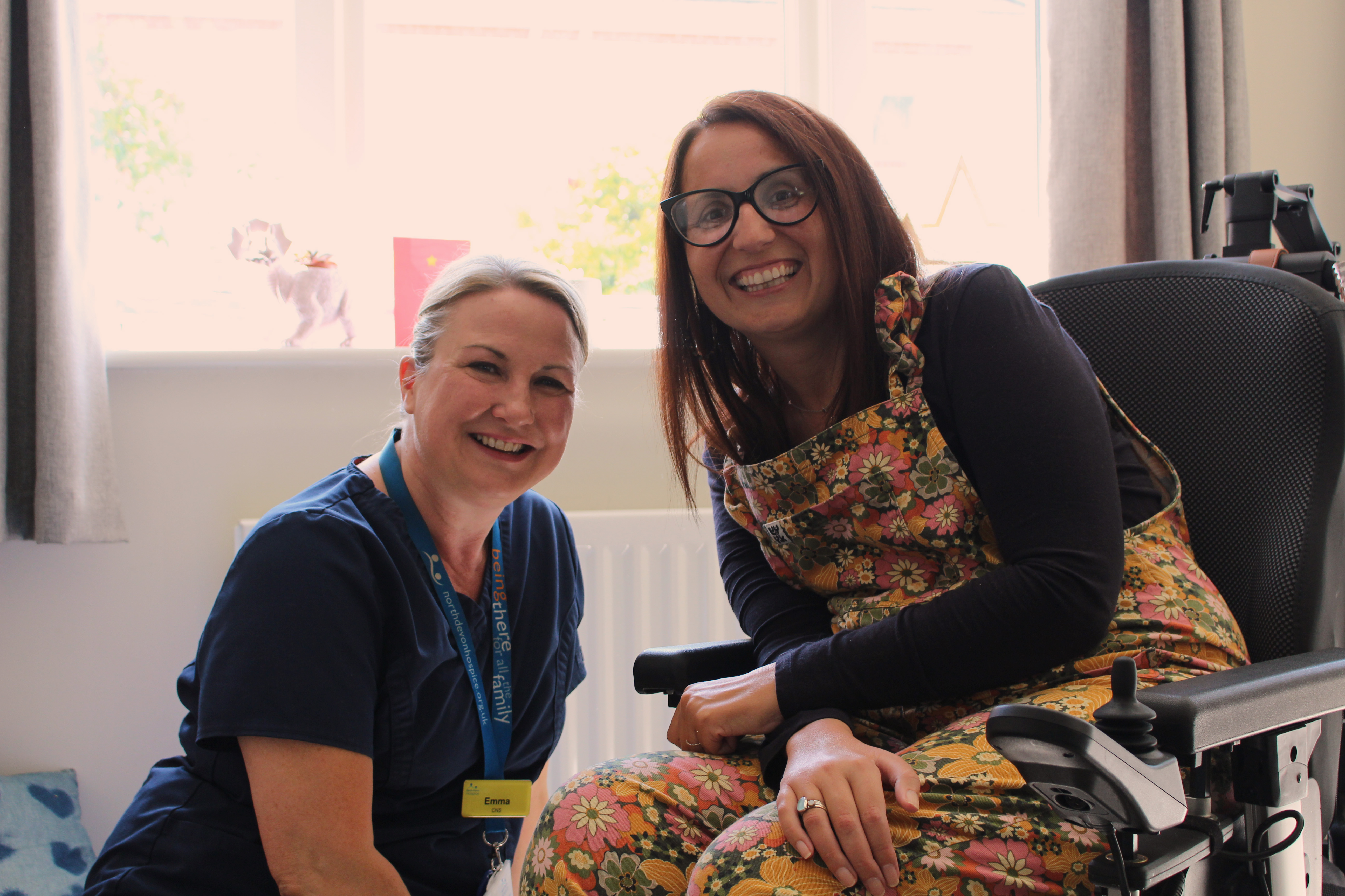 Female patient in a wheelchair sat with a female nurse in a lounge, smiling at the camera