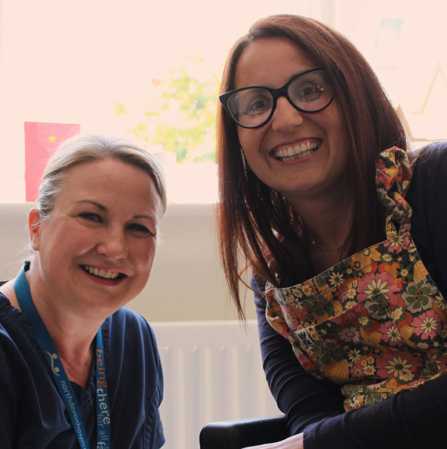Female patient in a wheelchair sat with a female nurse in a lounge, smiling at the camera