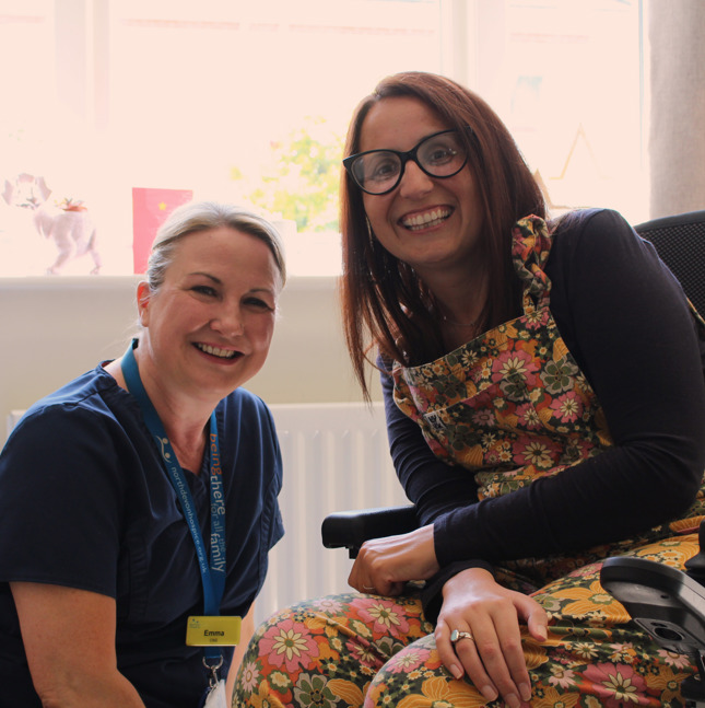 Female patient in a wheelchair sat with a female nurse in a lounge, smiling at the camera