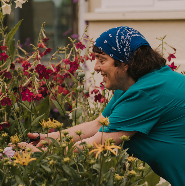 Female healthcare assistant in a teal tunic picking wild flowers from a garden, smiling