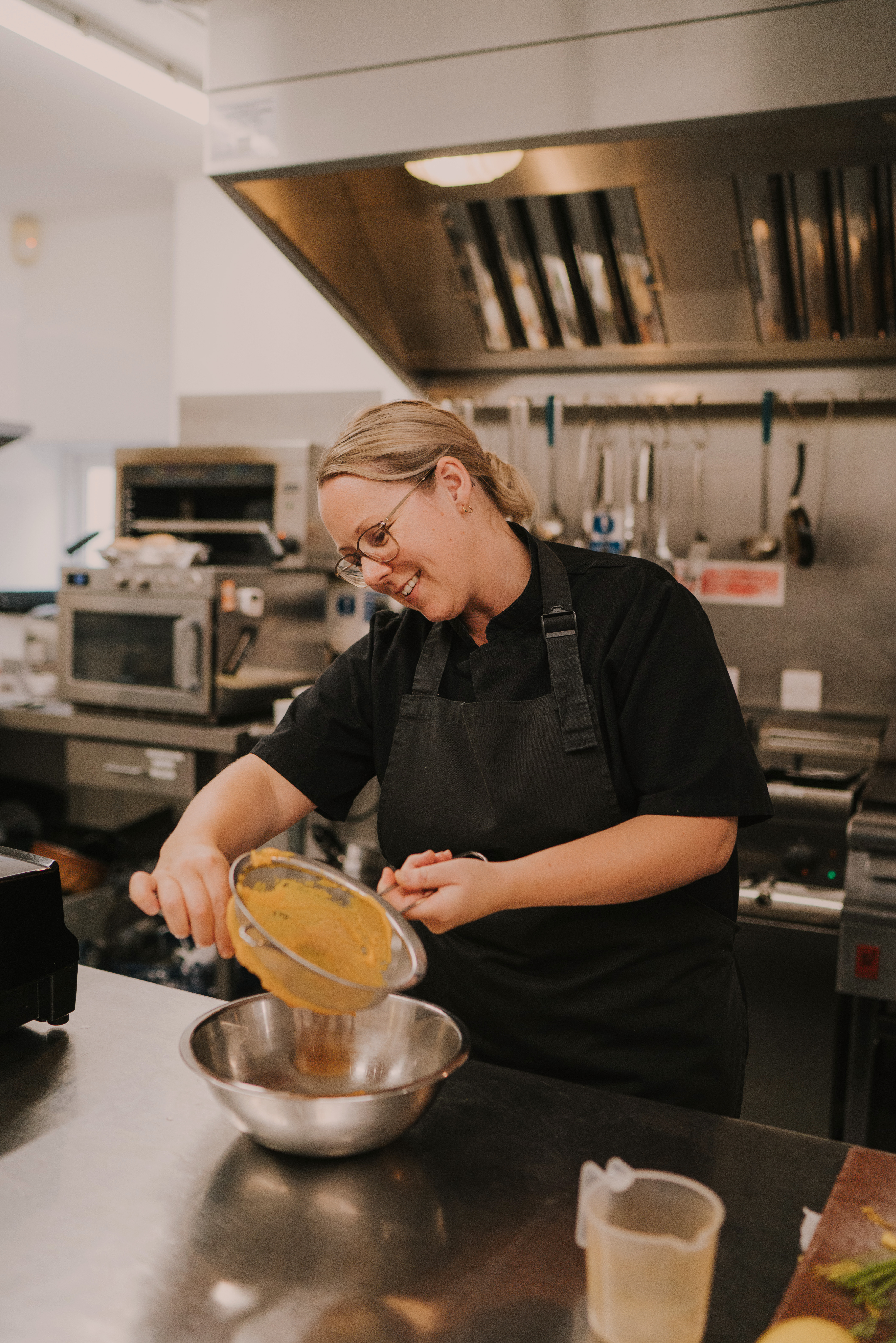 Female chef in black tunic, in an industrial kitchen, pushing puree through a sieve into a metal bowl