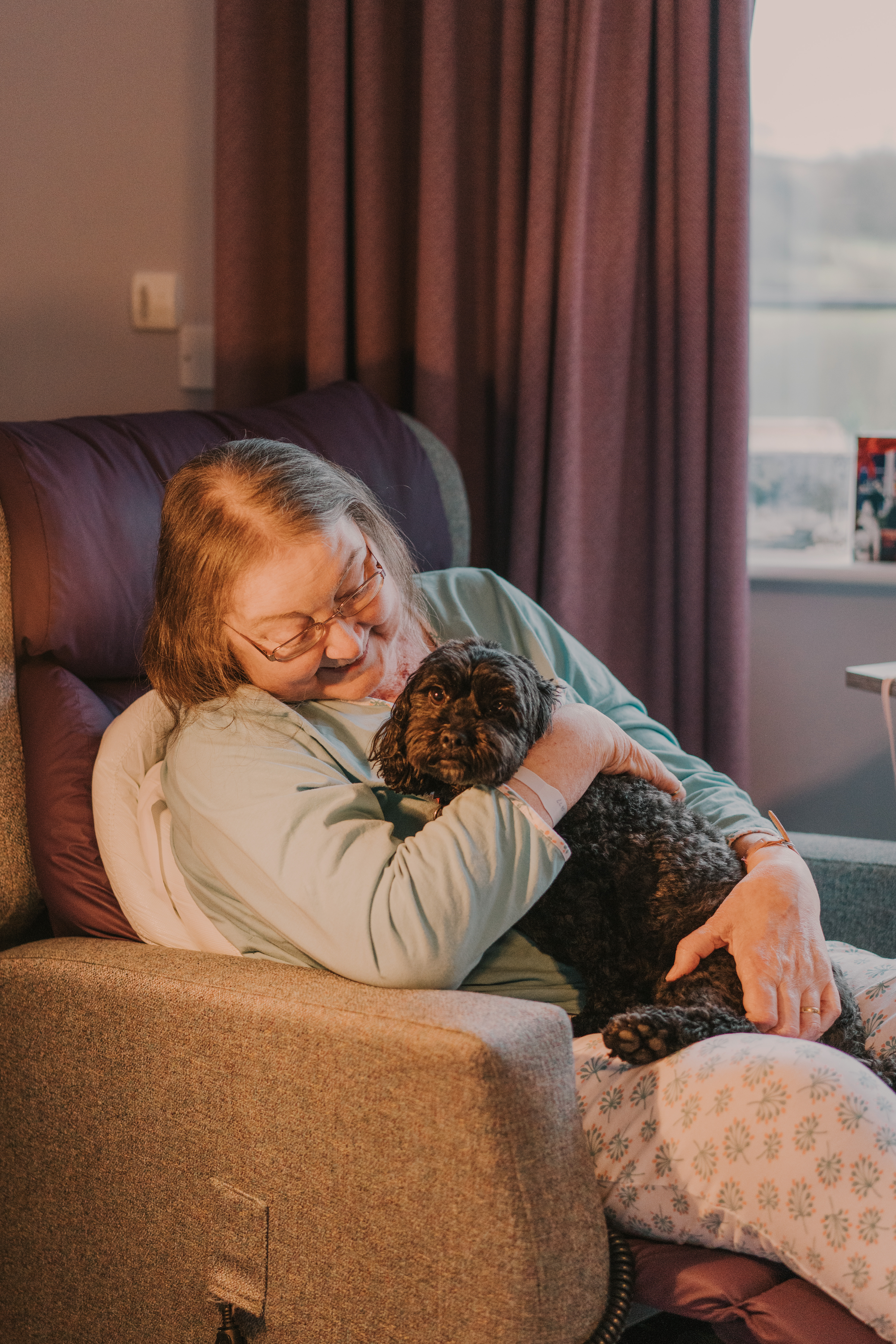 Female patient sitting in a grey chair holding a small, black therapy dog and smiling