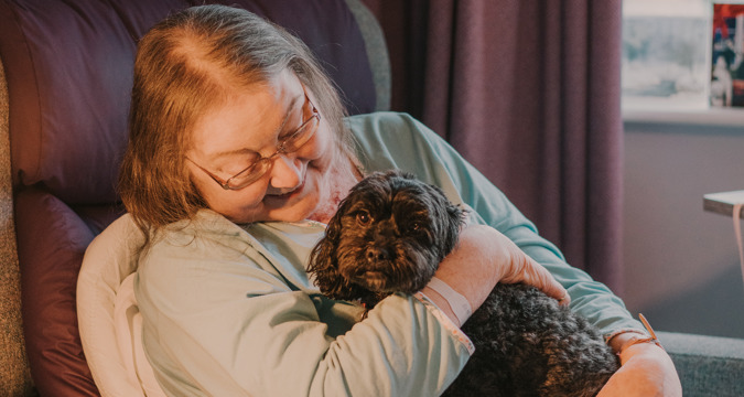 Female patient sitting in a grey chair holding a small, black therapy dog and smiling