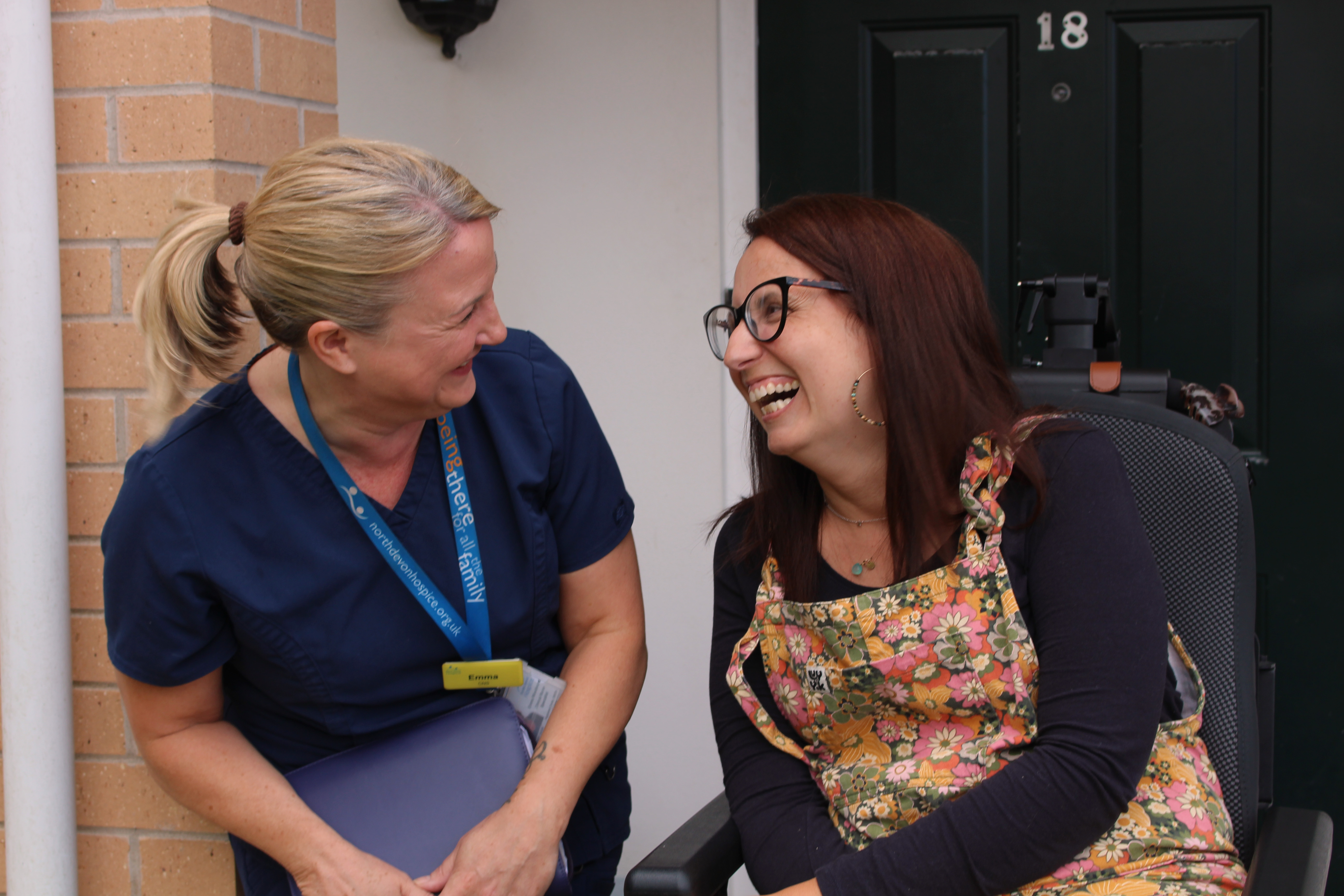 Female patient in a wheelchair smiling at a female nurse, outside a black front door with a number 18 on it