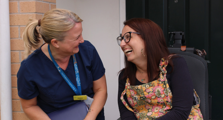 Female patient in a wheelchair smiling at a female nurse, outside a black front door with a number 18 on it