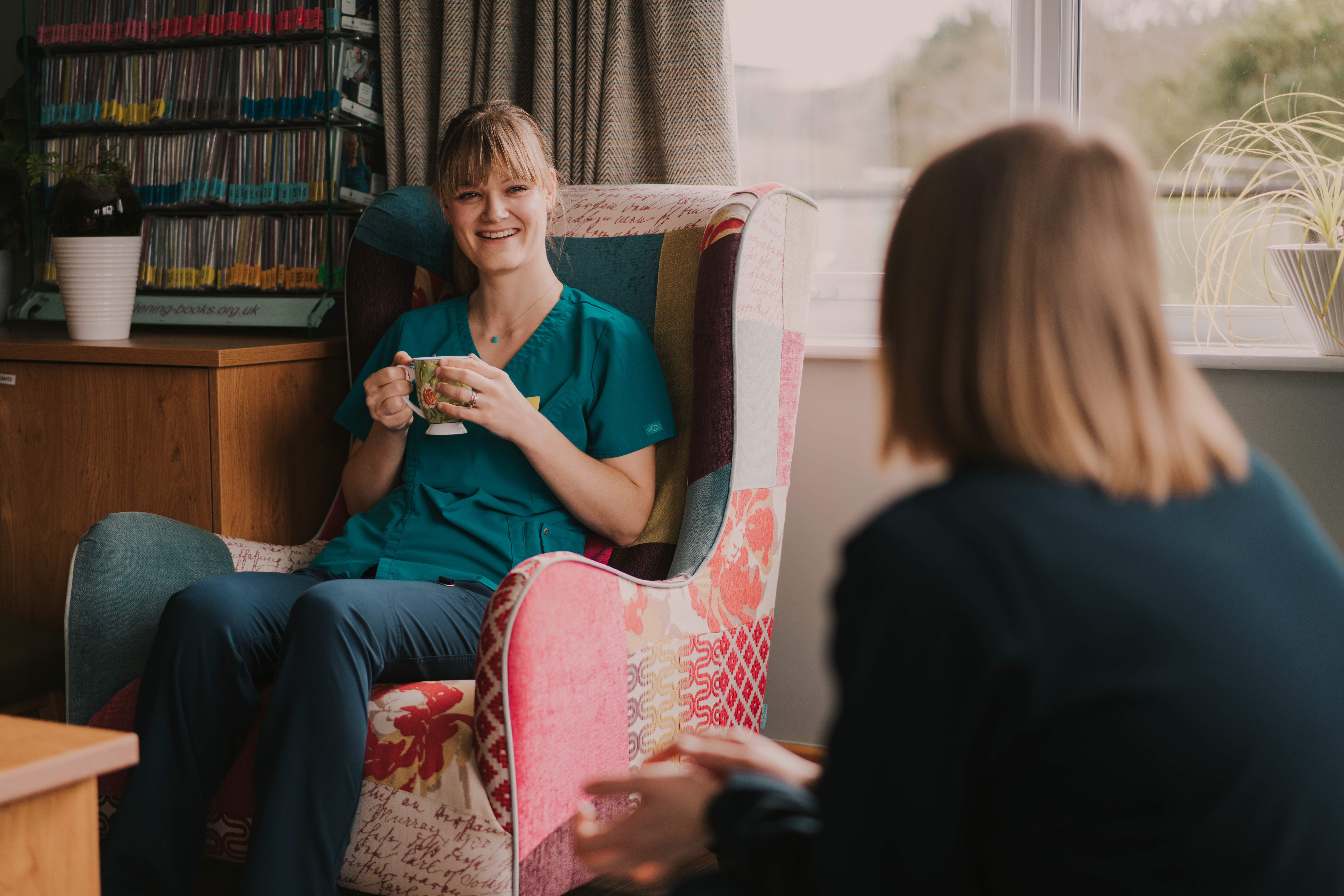 Female healthcare assistant sat on a patterned chair with a cup of tea, smiling at a female colleague