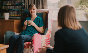 Female healthcare assistant sat on a patterned chair with a cup of tea, smiling at a female colleague