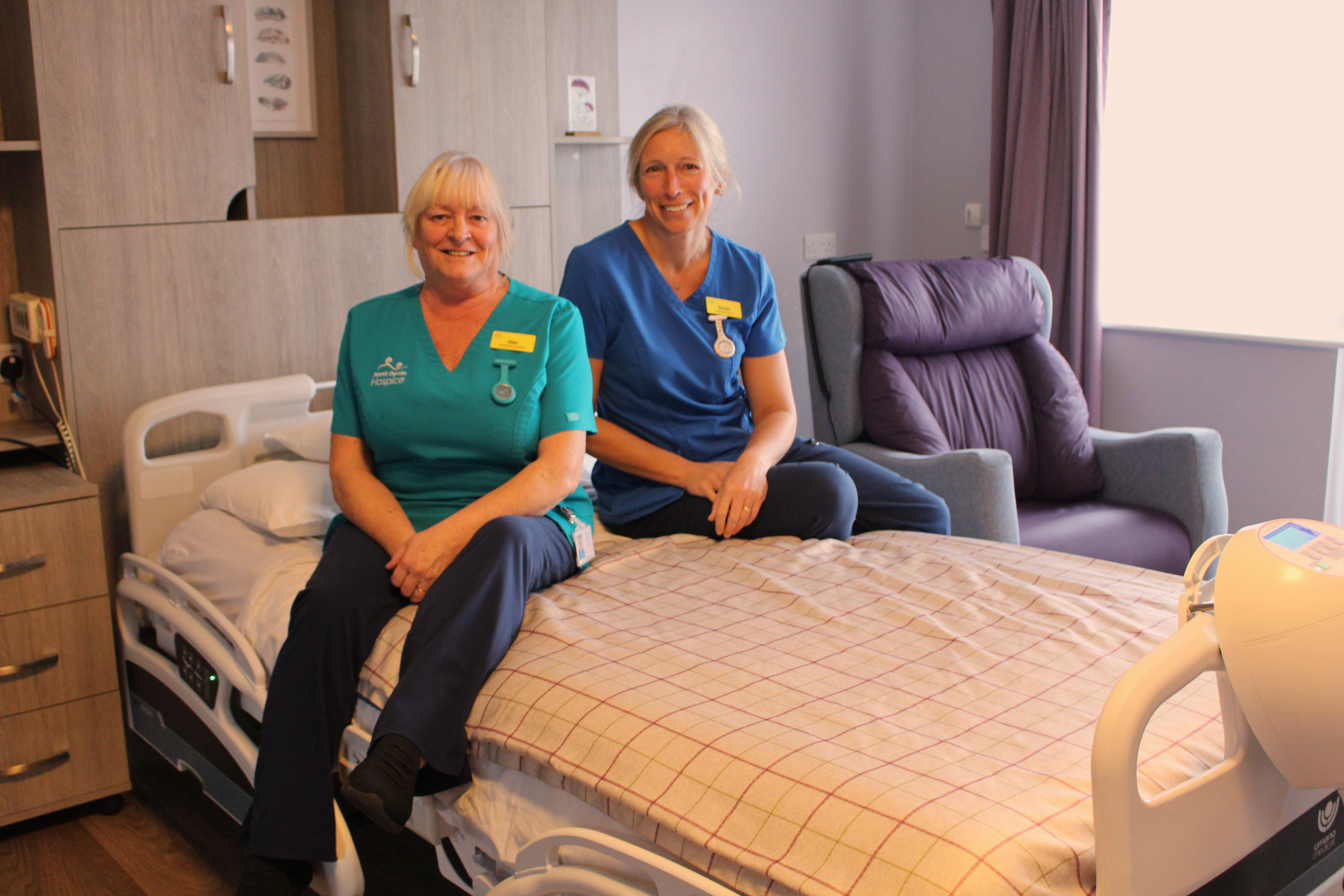 Image of two nurses, one in blue tunic and one in green tunic, sitting on a large bed in a patient bedroom with a chequered duvet cover