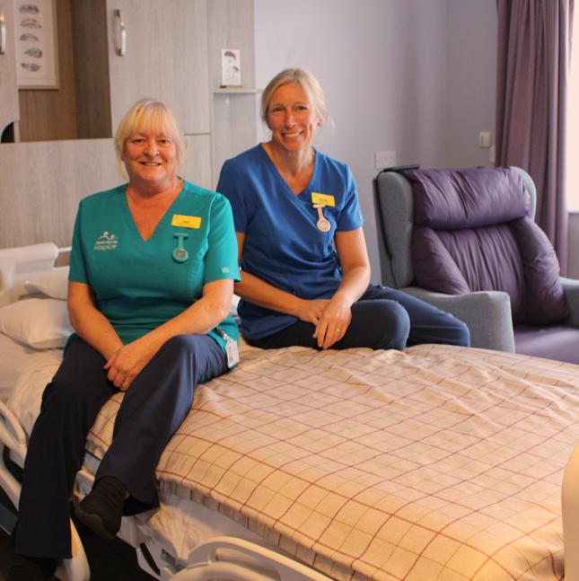 Image of two nurses, one in blue tunic and one in green tunic, sitting on a large bed in a patient bedroom with a chequered duvet cover