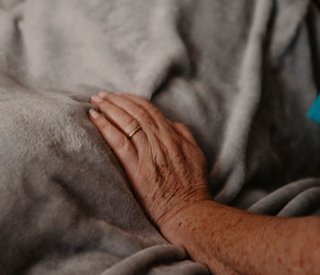 Female healthcare assistant, with her hand resting on a grey blanket of a patient in a hospital bed