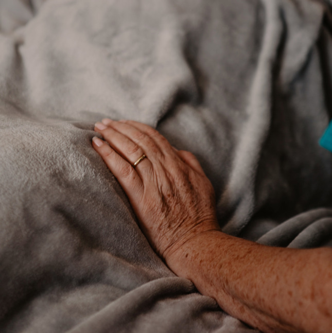 Female healthcare assistant, with her hand resting on a grey blanket of a patient in a hospital bed