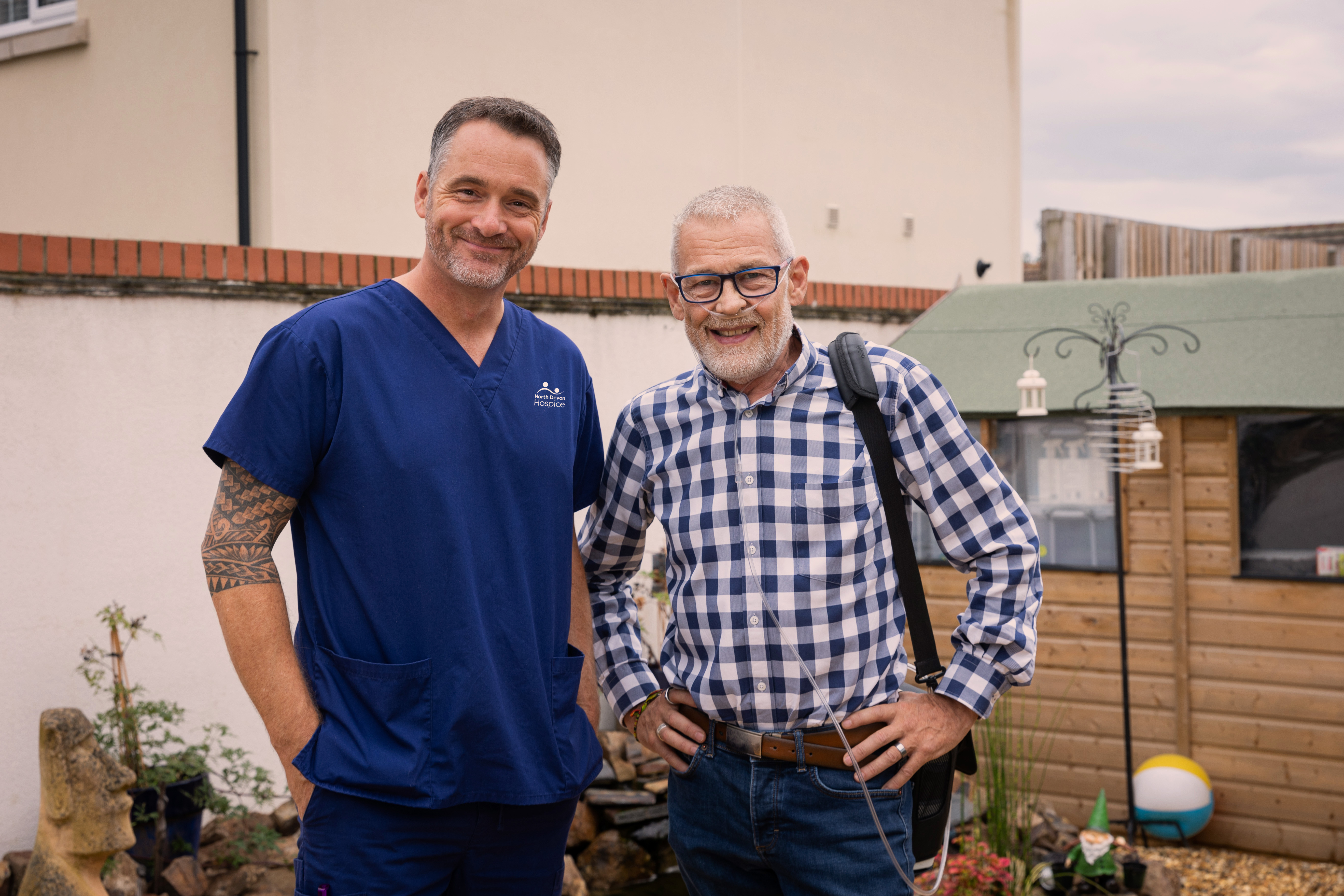 Male patient with oxygen tube, stood with male nurse in a back garden, smiling at the camera