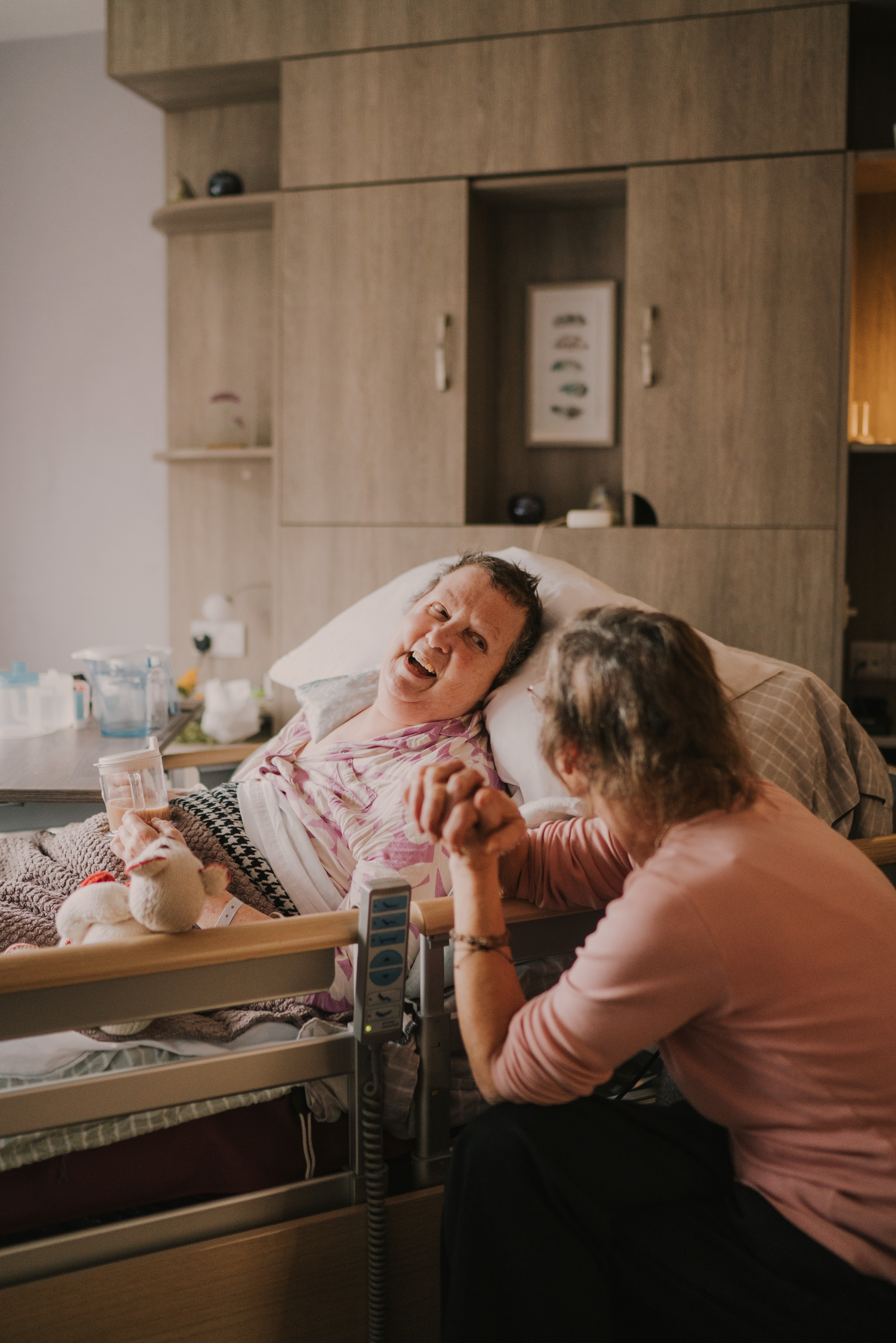 Female patient in a hospital bed, drinking tea from a cup while smiling at a female family member to her left