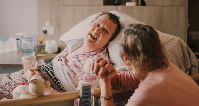 Female patient in a hospital bed, drinking tea from a cup while smiling at a female family member to her left