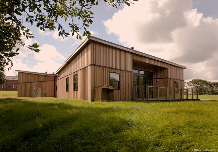 Image of a large wooden, square building surrounded with grass, with sunshine and clouds in the sky