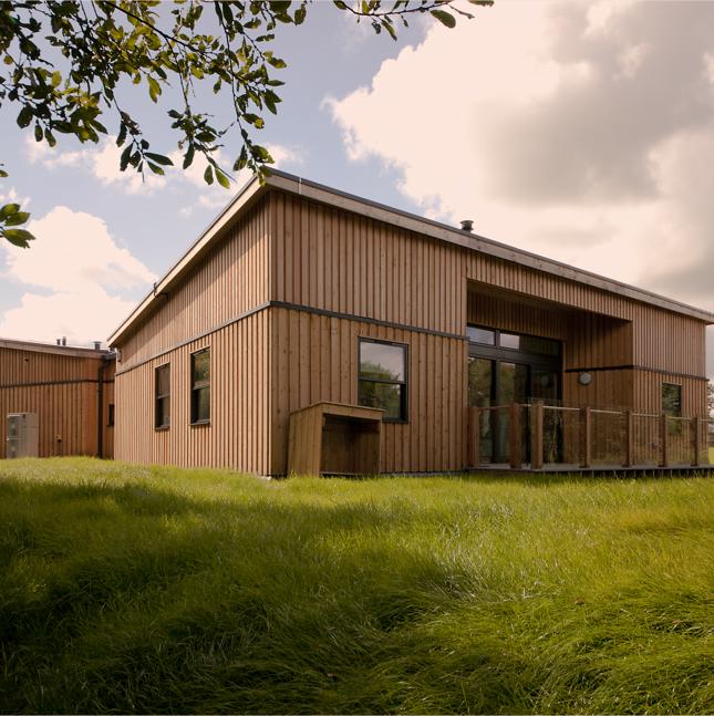Image of a large wooden, square building surrounded with grass, with sunshine and clouds in the sky
