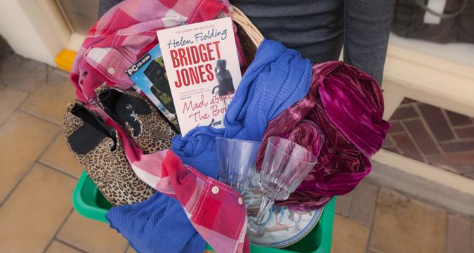 Female in a grey top holding a green box filled with donations - wine glasses, leopard print boots, books and clothing
