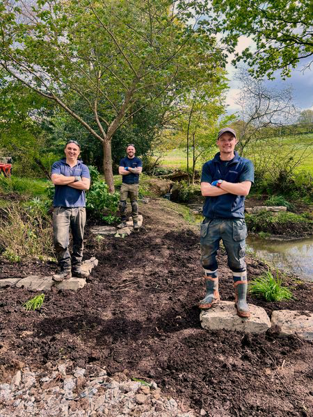 Three males stood in a garden they have been working on, with arms folded and smiles on their faces