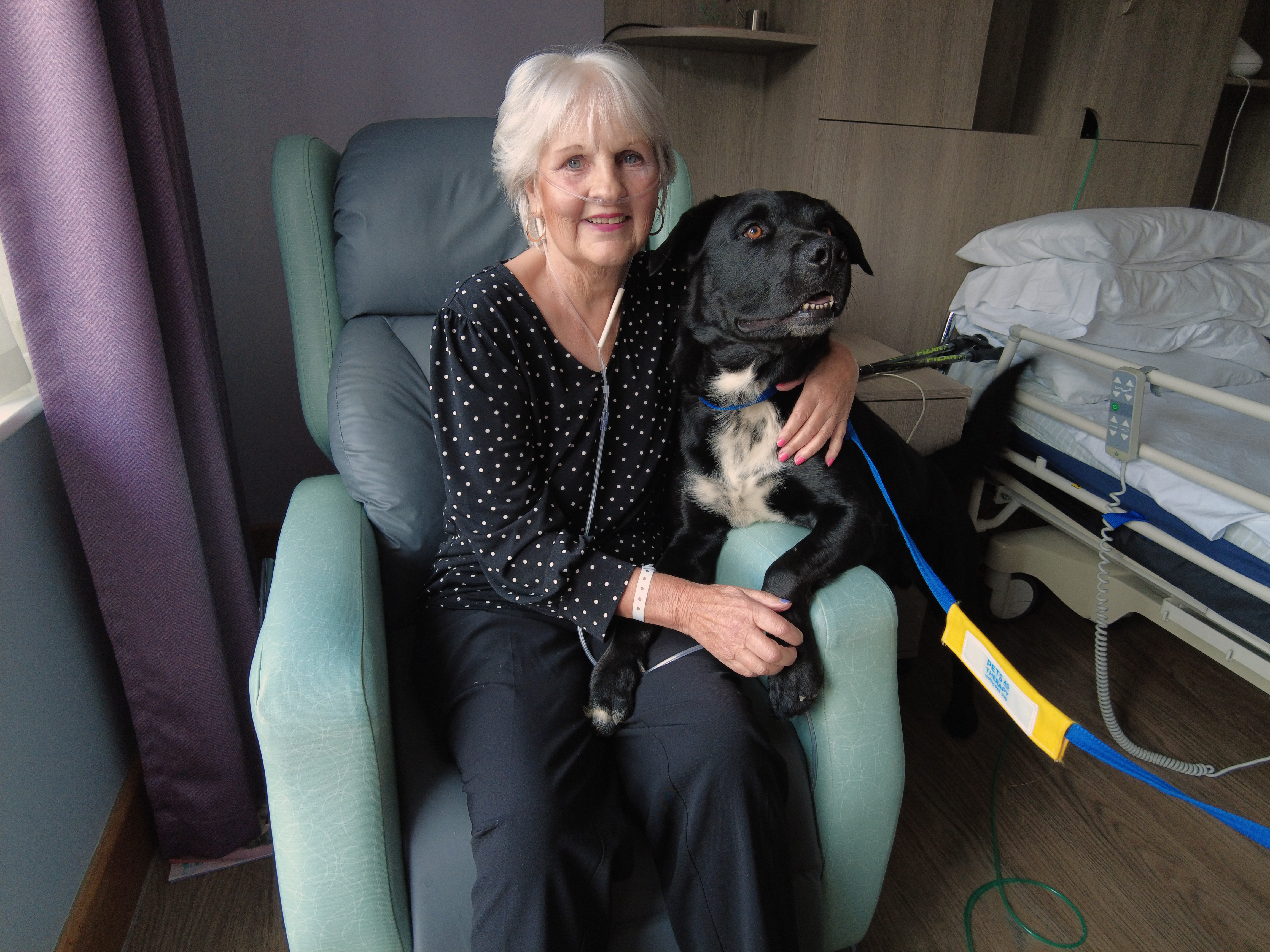 Female patient in a green chair cuddling a large, black therapy dog, smiling