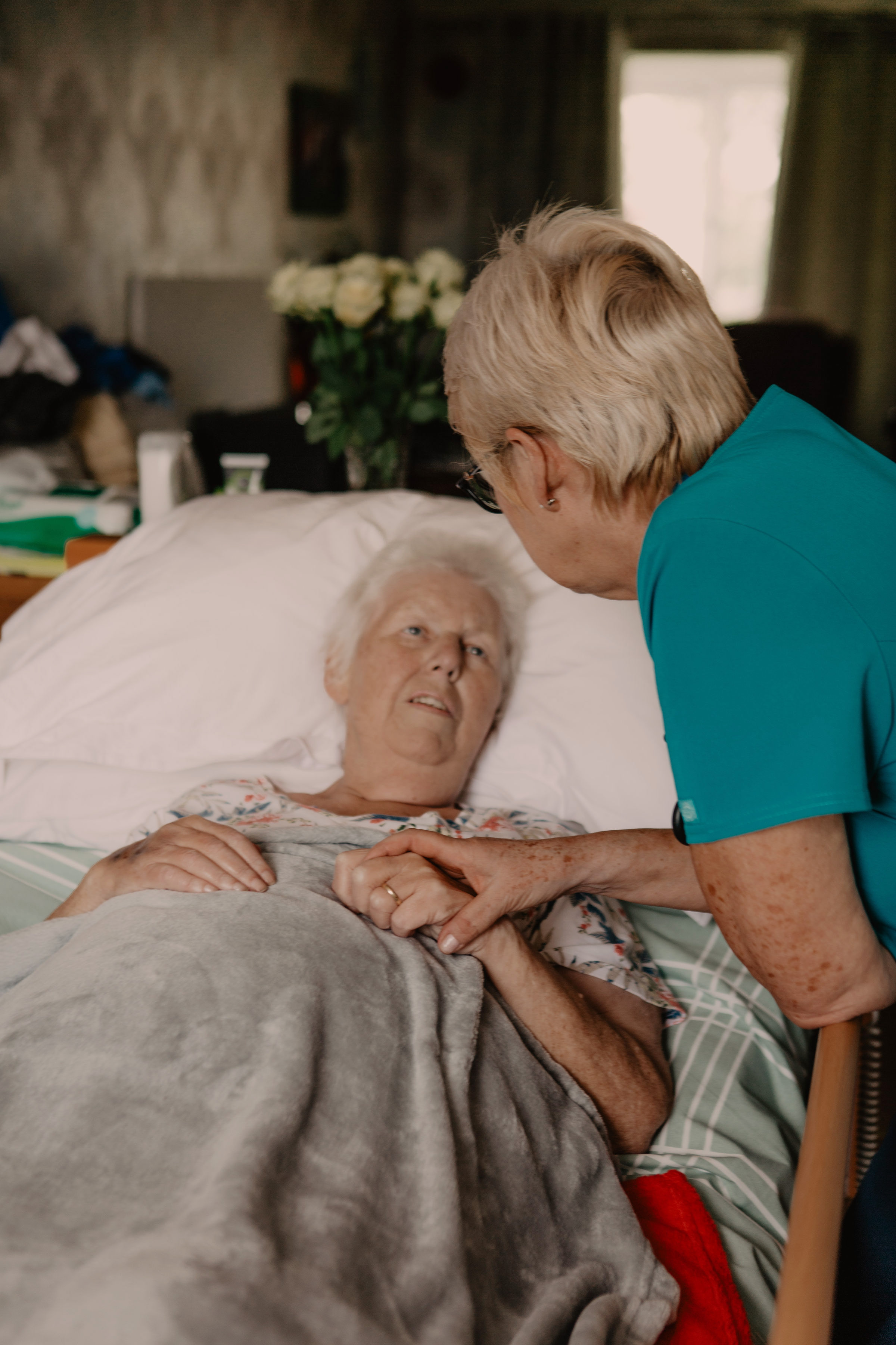 Female healthcare assistant, holding the hand of a female patient lying in a hospital bed in her home