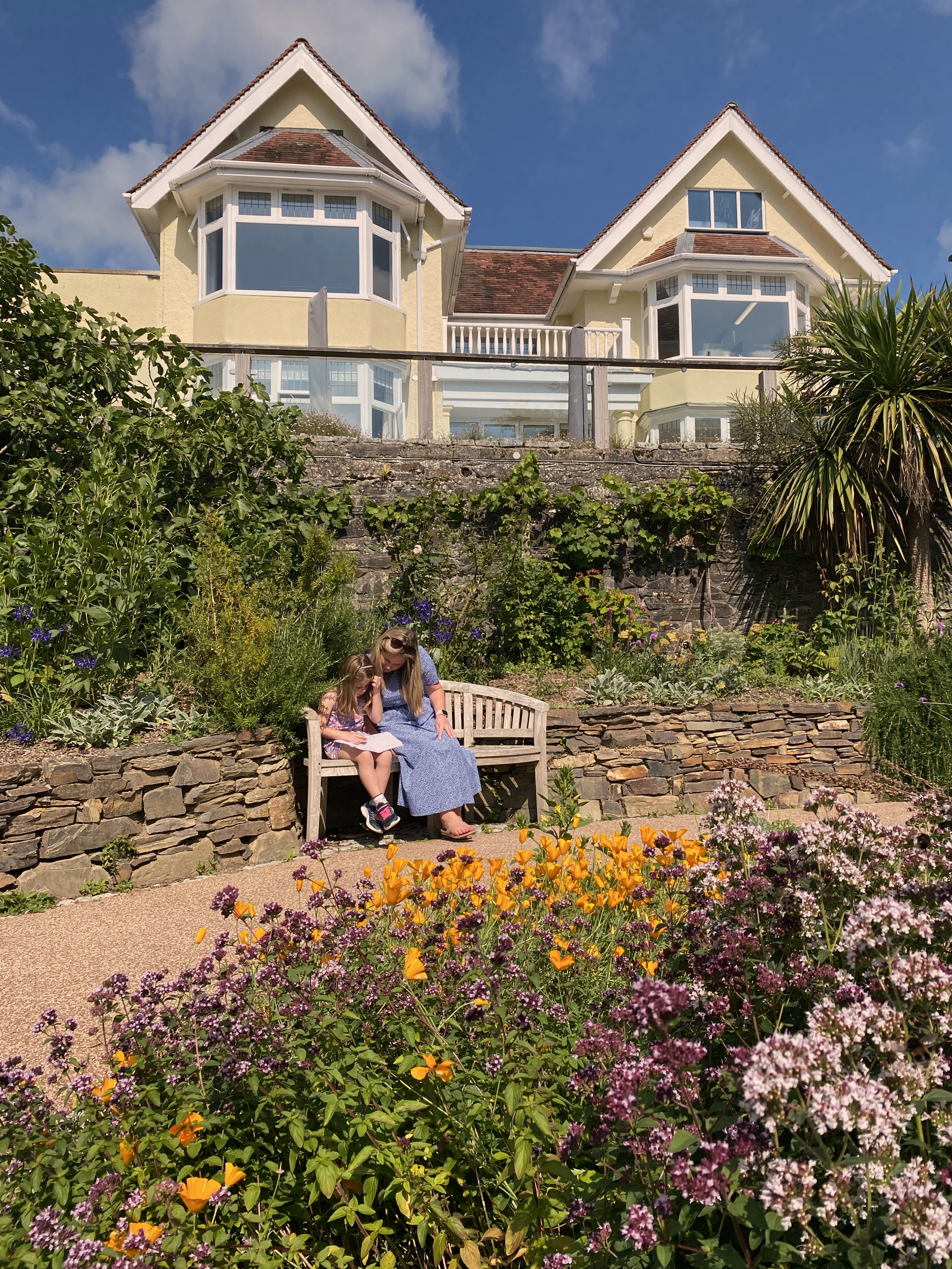 Female in a blue dress sat on a bench next to a small female child, surrounded by flowers and the hospice in the background