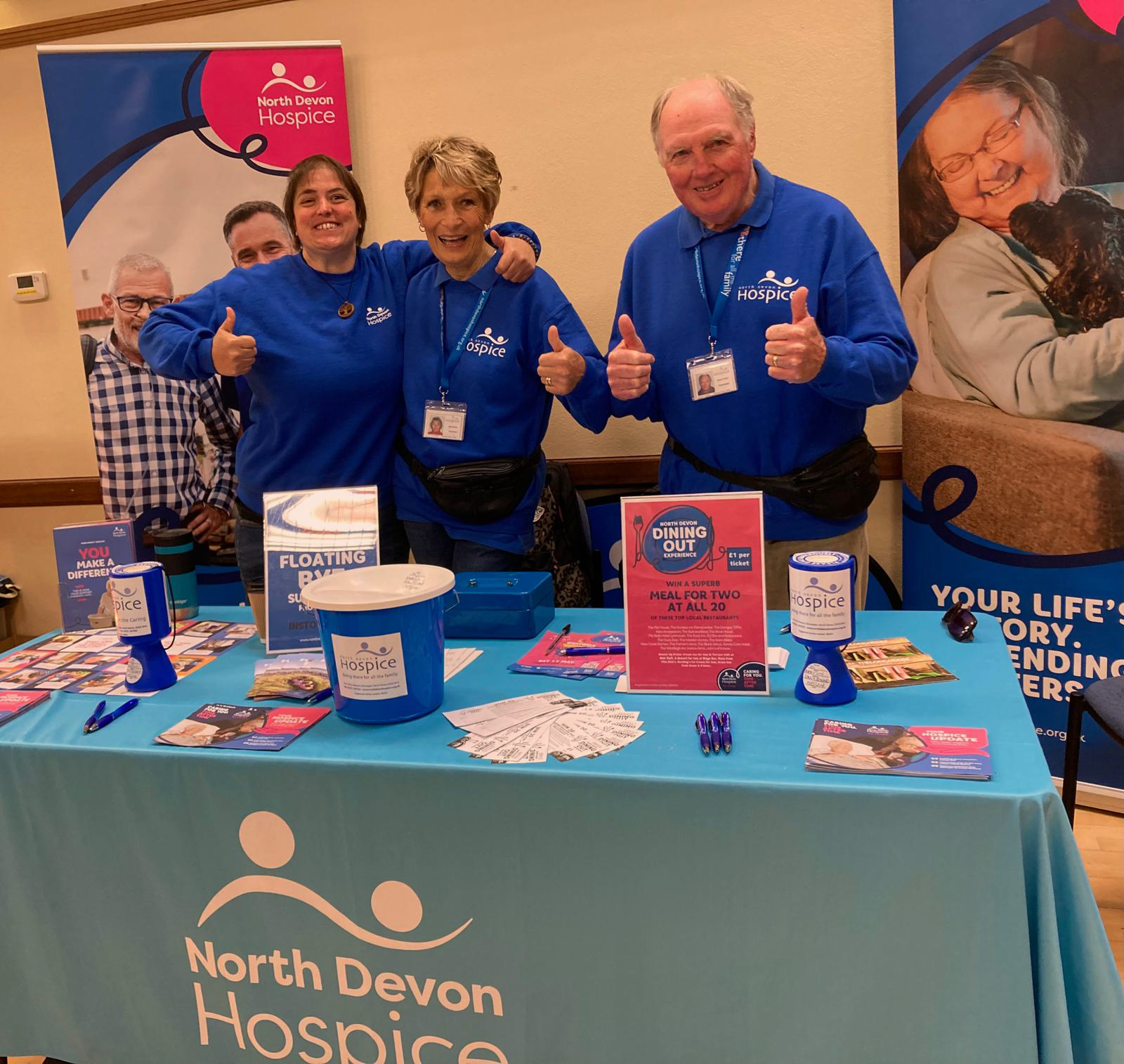 Two females and a male in blue hospice branded jumpers, stood behind table full of branded hospice items, with thumbs up and facing camera smiling