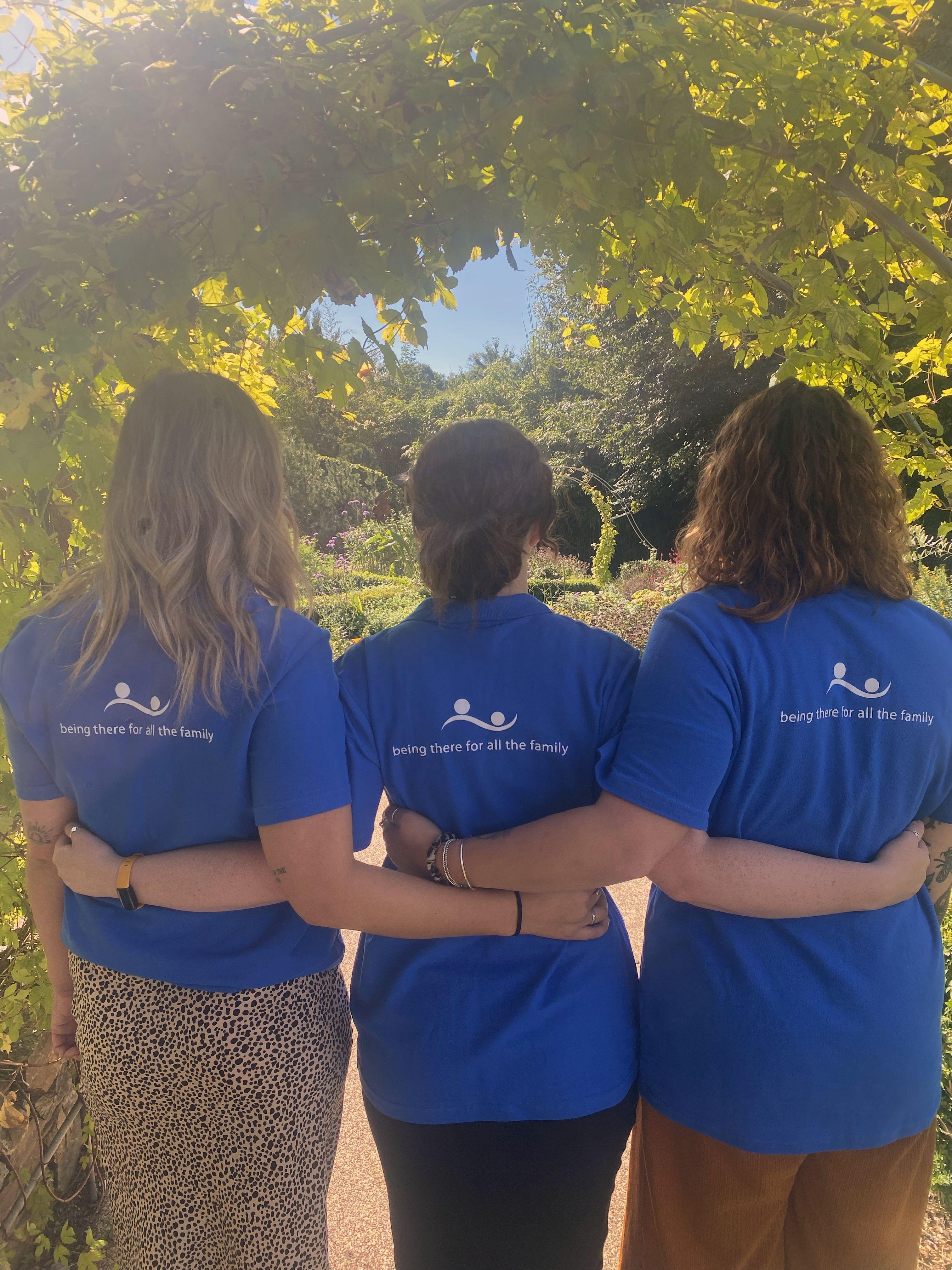 Three females stood facing away from the camera, wearing blue polos with the hospice logo on the back, arms round each other in the garden in summer