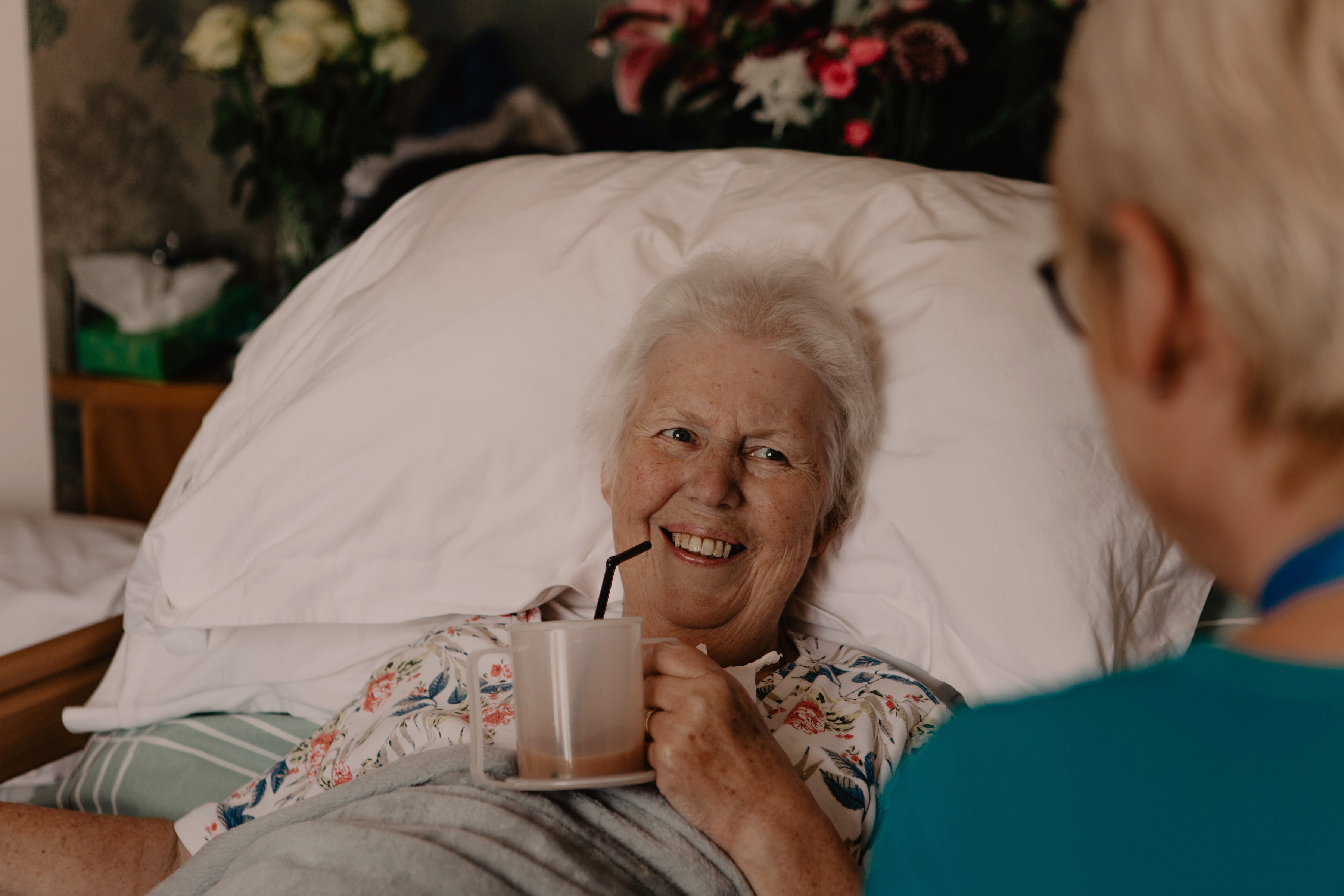 Female patient in a hospital bed, drinking tea from a cup while smiling at a female healthcare assistant
