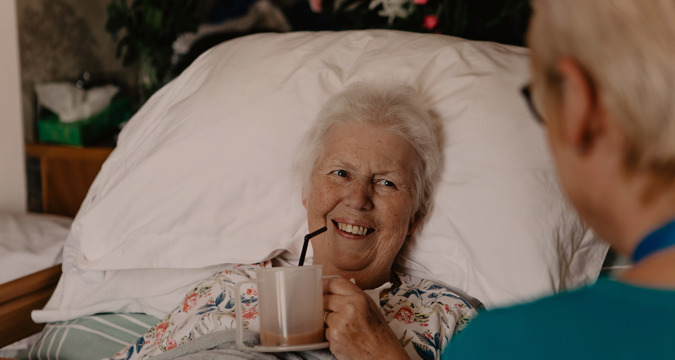 Female patient in a hospital bed, drinking tea from a cup while smiling at a female healthcare assistant