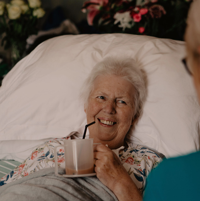 Female patient in a hospital bed, drinking tea from a cup while smiling at a female healthcare assistant
