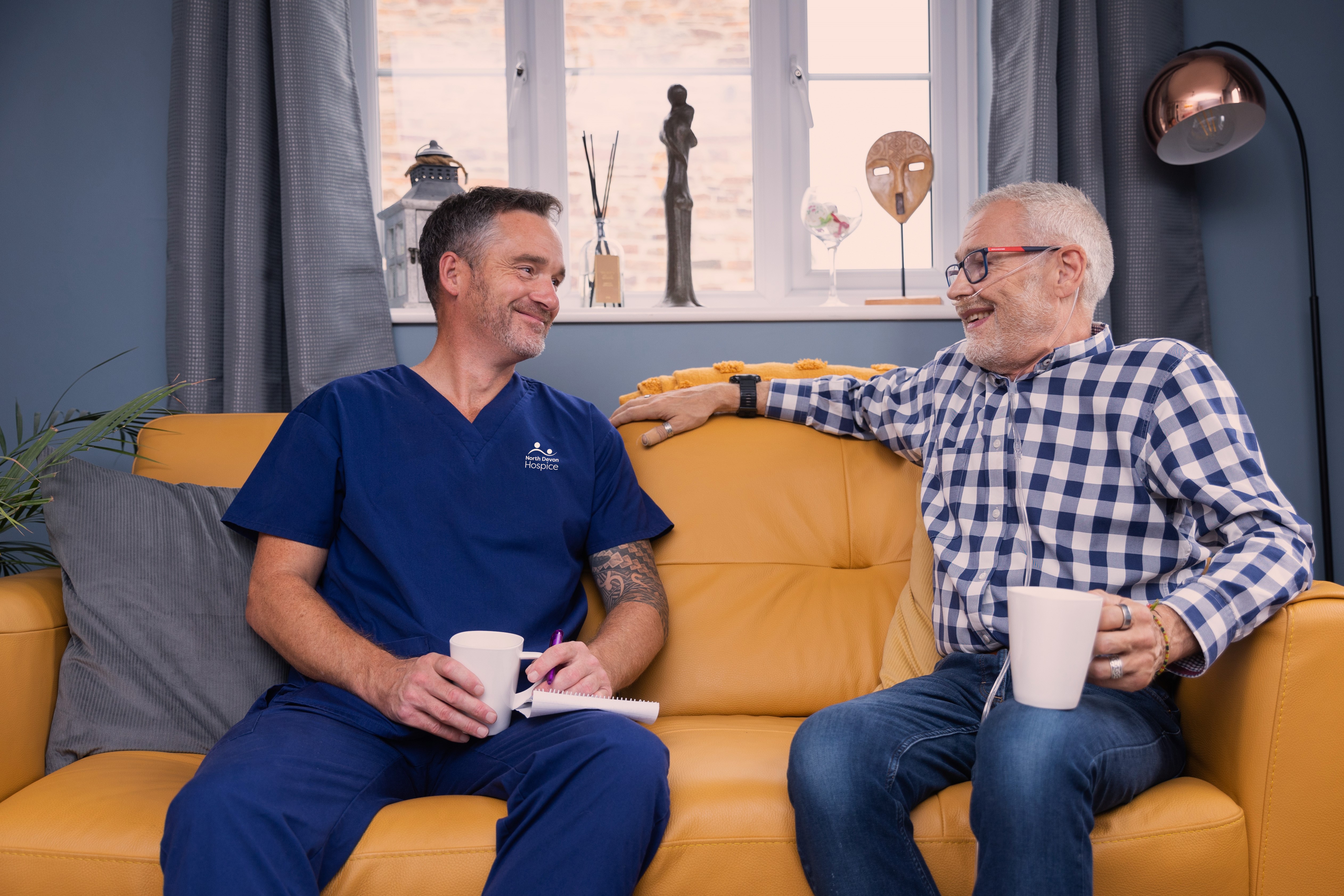 Male patient sat on a yellow sofa with male nurse, drinking cups of tea and smiling at each other