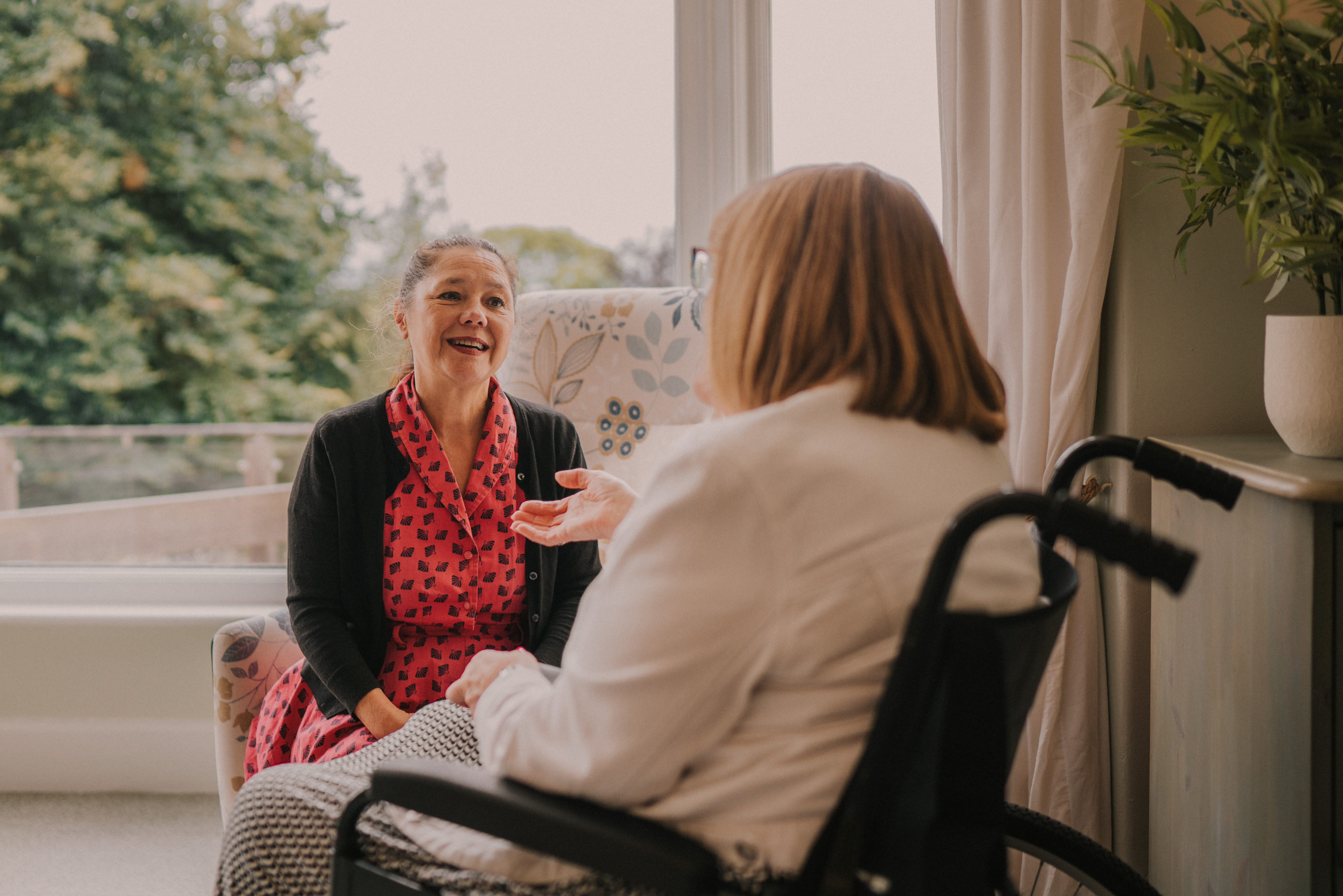 Female patient sat in a wheelchair having a conversation with a female counsellor with a window in the background