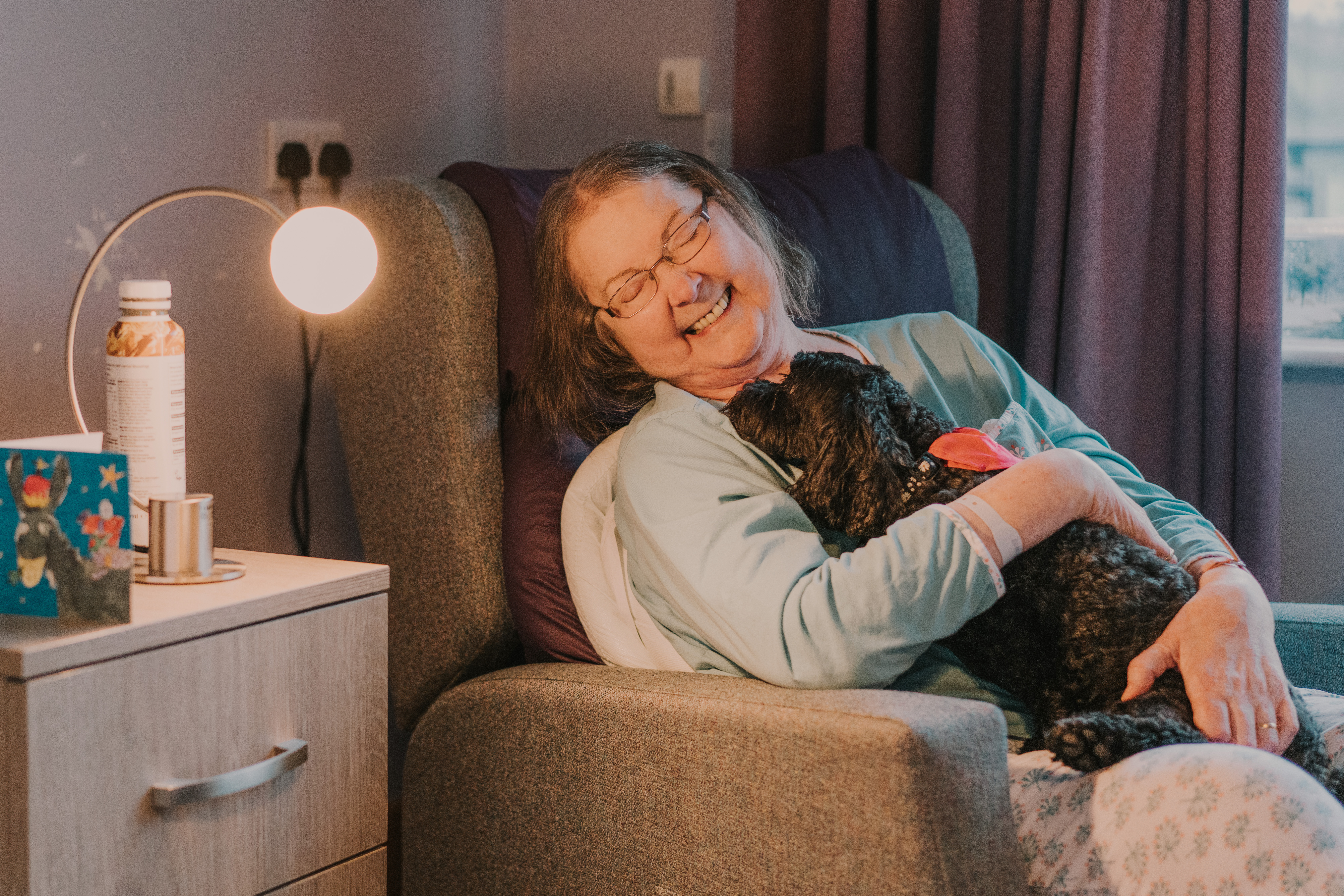 Patient seated in a hospice bedroom, smiling while cuddling a small, black therapy dog