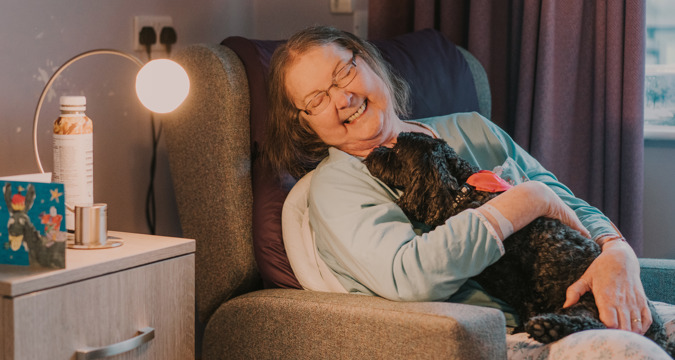 Patient seated in a hospice bedroom, smiling while cuddling a small, black therapy dog